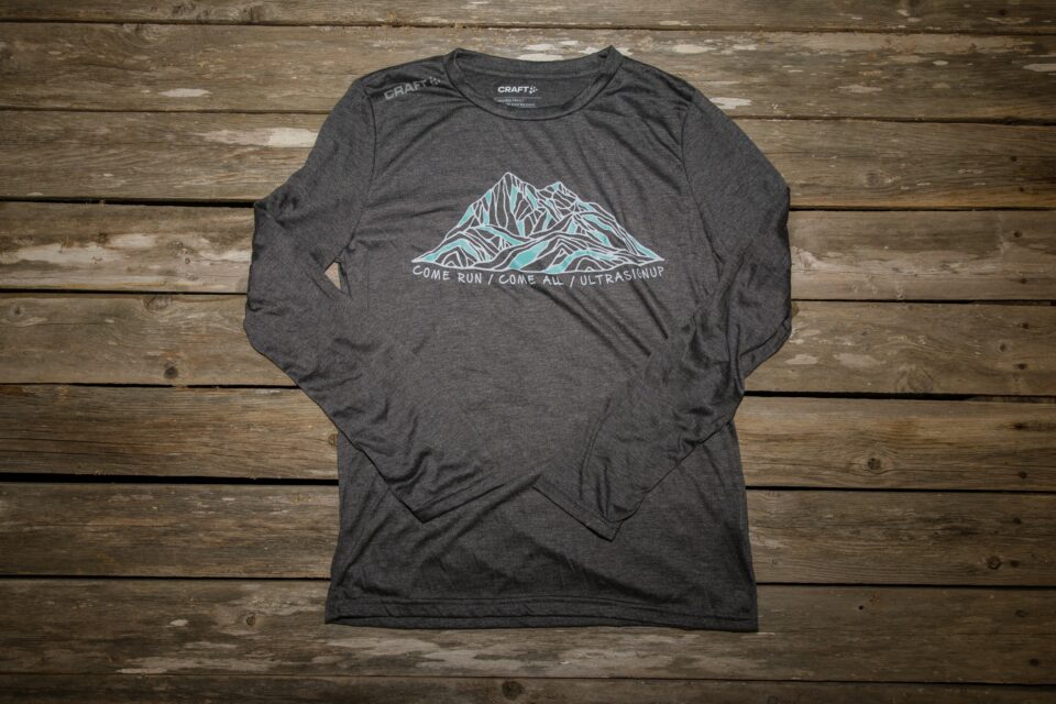 Gray long-sleeve crew neck t-shirt lying face up on a background of wooden slats. The t-shirt has a design across the chest. The design is a fine-line drawing of a mountain in white and blue lines. Underneath the design are the words "Come Run / Come All / UltraSignup." The right shoulder says "Craft."