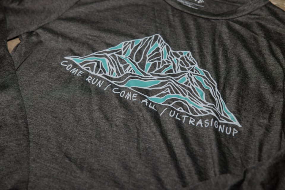 A close up view of a design on the chest of a gray t-shirt. The design is a fine-line drawing of a mountain in white and blue lines. Underneath the design are the words "Come Run / Come All / UltraSignup."