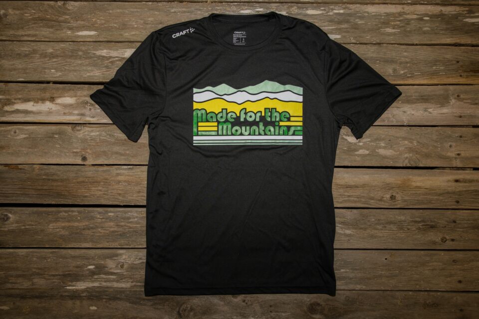 Black crew neck short sleeve t-shirt lying face up on a background of wooden slats. The t-shirt has a design across the chest. The design is made up of tri-color white, green, and yellow thick lines that are interrupted by green text that says "made for the mountains" and above the text the lines turn into mountains shaded in white, green, and yellow. The right shoulder says "Craft."