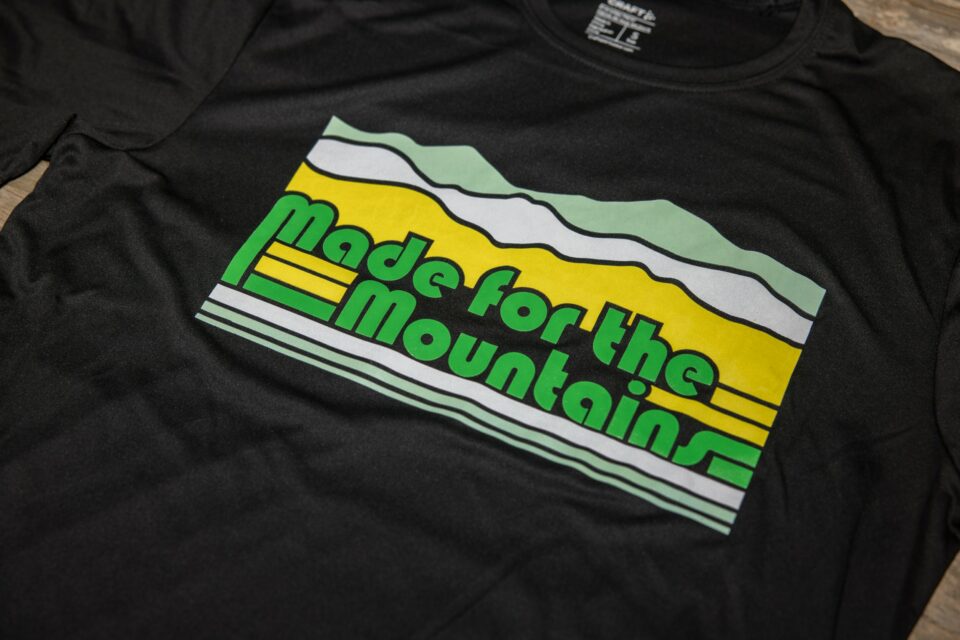 A close up view of a design on the chest of a black t-shirt. The design is made up of tri-color white, green, and yellow thick lines that are interrupted by green text that says "made for the mountains" and above the text the lines turn into mountains shaded in white, green, and yellow.